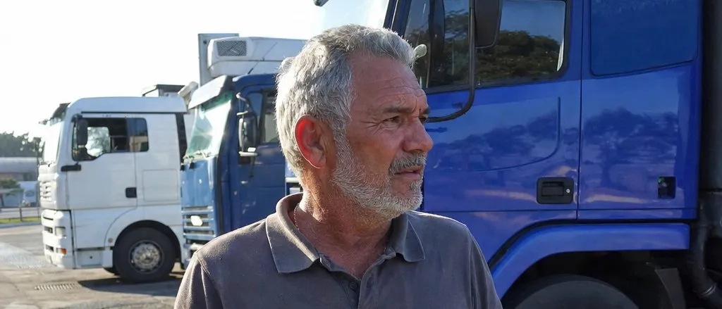 Caminhoneiro de cabelo grisalho e barba branca em pé no pátio de um posto, com carretas estacionadas atrás no fim da tarde