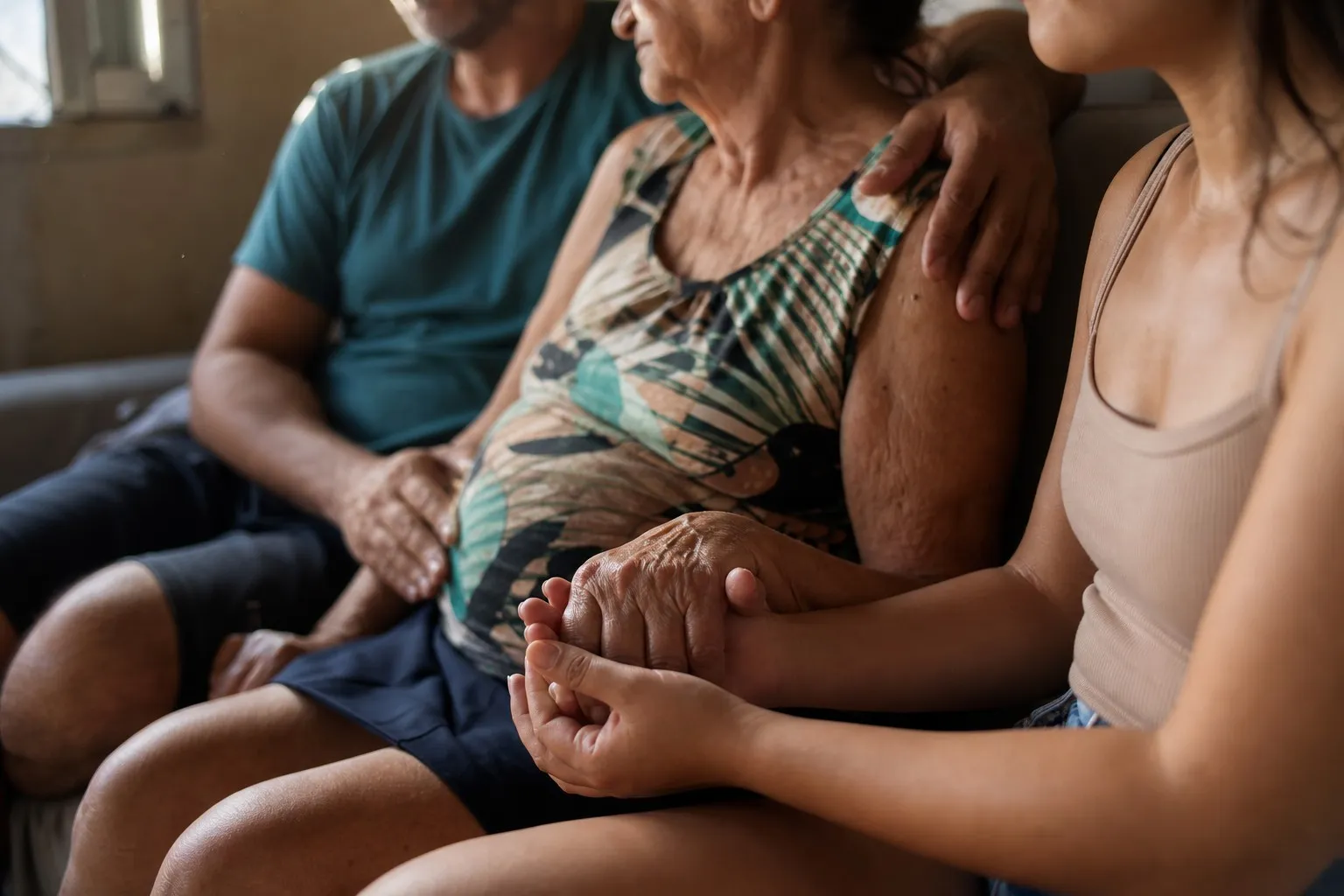 Três pessoas de idades diferentes sentadas lado a lado num sofá, enquadradas do pescoço para baixo. Uma mulher mais velha está no meio, com a mão de uma jovem segurando a dela no colo e o braço de um homem apoiado no ombro dela