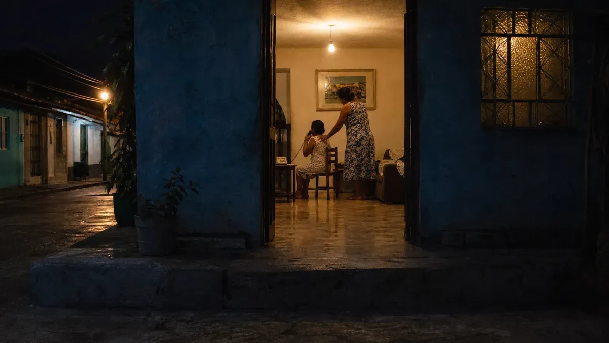 Casa simples de rua à noite, com o chão molhado de chuva, vista de fora pela porta aberta. Na sala iluminada por uma lâmpada, uma mulher está sentada numa cadeira com um telefone fixo no ouvido, e outra mulher em pé mantém a mão apoiada no ombro dela