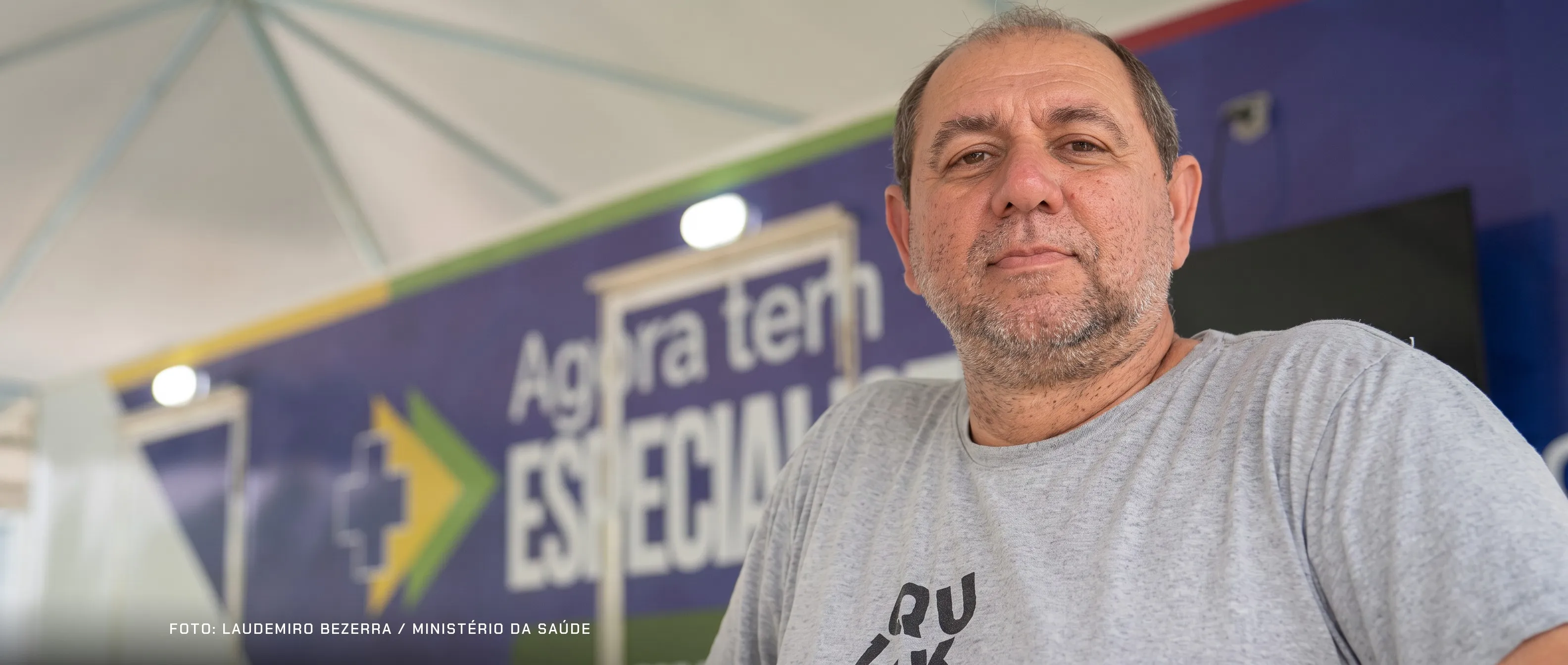 Caminhoneiro brasileiro de cerca de 50 anos, cabelo grisalho e barba por fazer, de camiseta cinza, em pé dentro da tenda de uma unidade de saúde do SUS instalada num ponto de parada. Atrás dele, desfocado, o painel do programa Agora Tem Especialistas do Ministério da Saúde, com a cruz e a seta nas cores da bandeira